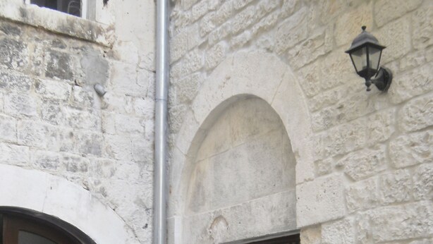 A stone building with two wooden doors, a small entrance, and a potted plant with pink flowers. - Palace Stafileo (Trogir)