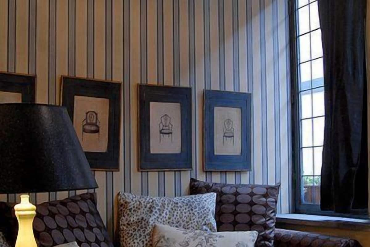 Ein Schlafzimmer mit gestreifter Tapete, einem Bett mit gemusterten Betttextilien, einem Nachttisch mit Lampe und gerahmten Bildern an der Wand.