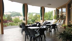 An outdoor patio area with tables and chairs, surrounded by potted plants and large windows offering a view of the landscape. - Hotel Ducale (Salsomaggiore Terme)