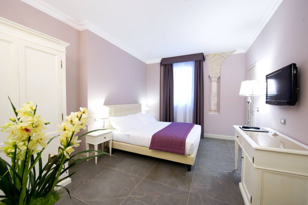 Une chambre d’hôtel comprenant un lit, une table de chevet, une télévision, une salle de bain avec un miroir et une fenêtre avec des rideaux.