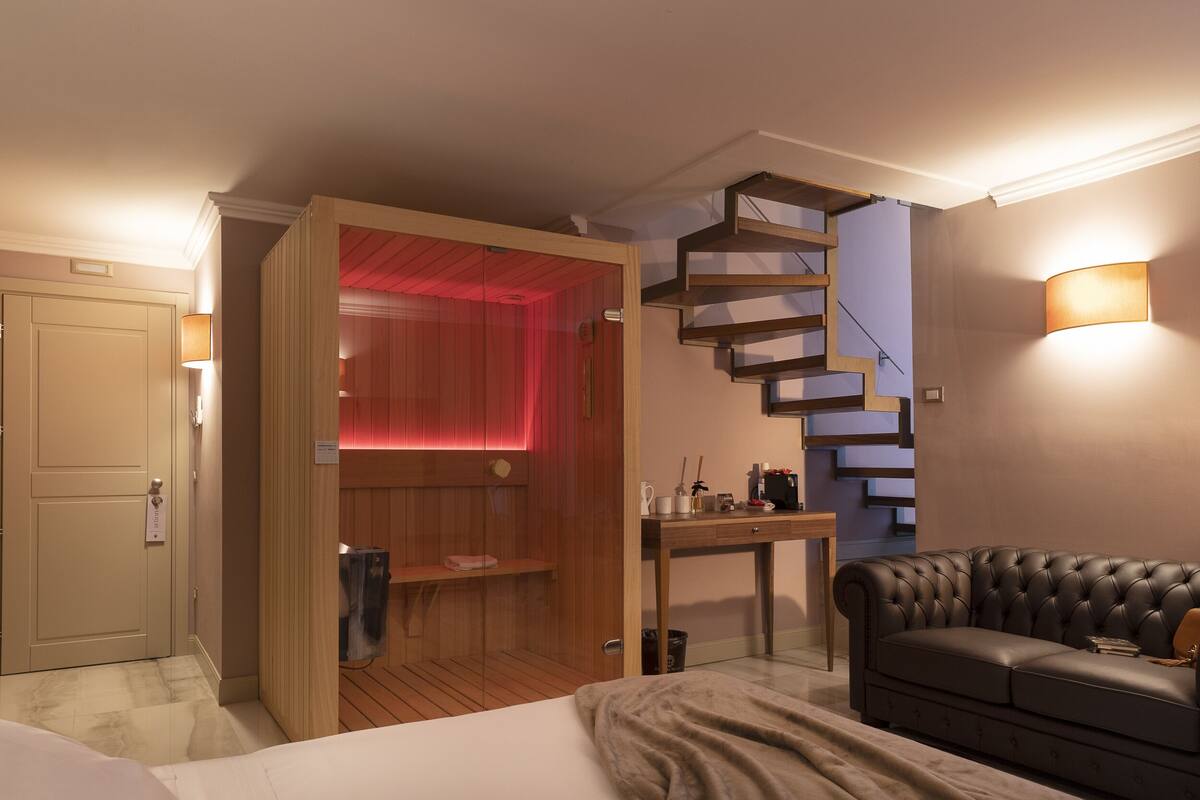Une chambre d’hôtel avec un lit, un bureau en bois, un canapé en cuir noir, un sauna éclairé de lumières rouges et un escalier en colimaçon.