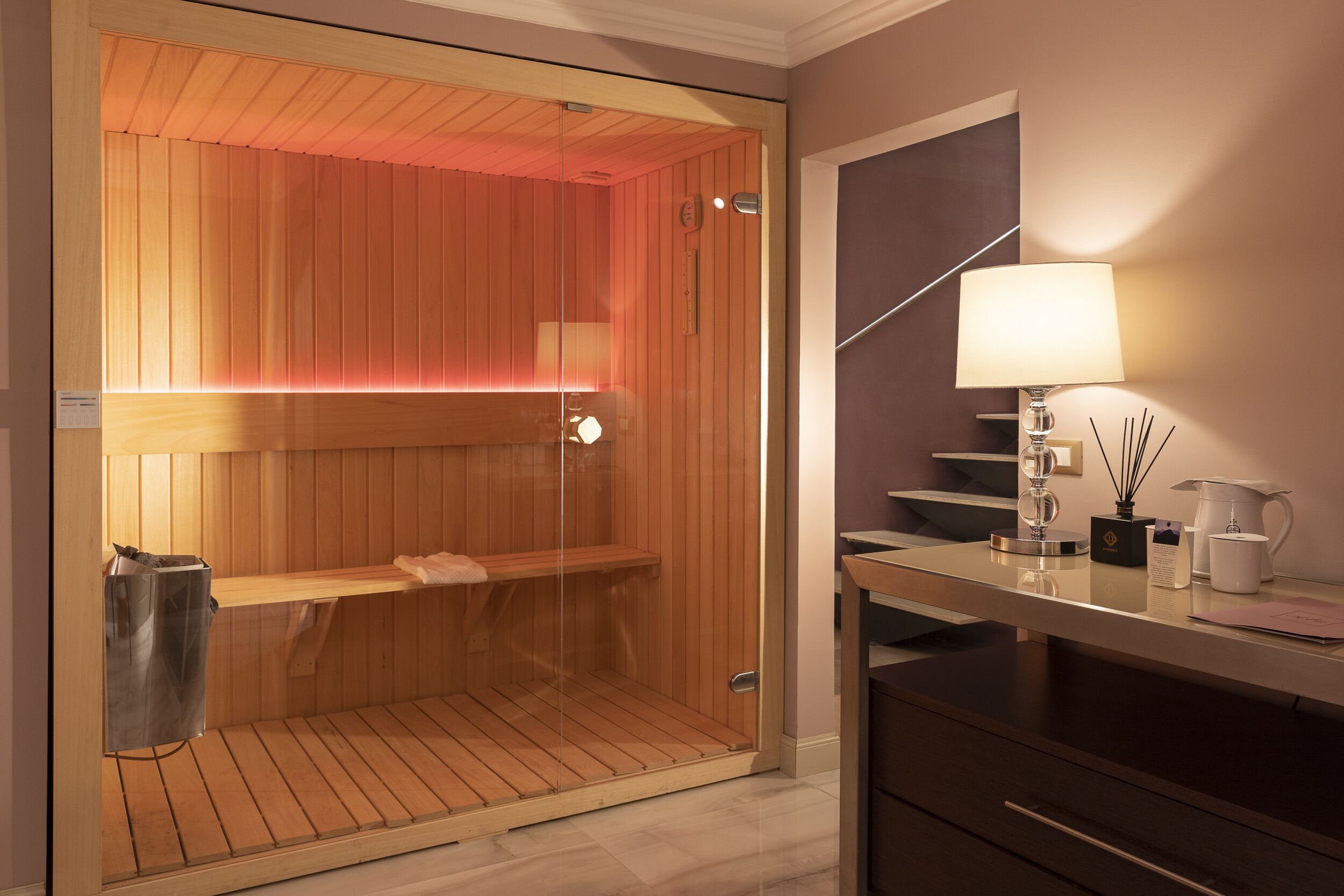 A sauna with a wooden interior and a glass door, a white lamp, and a coffee maker on a counter.