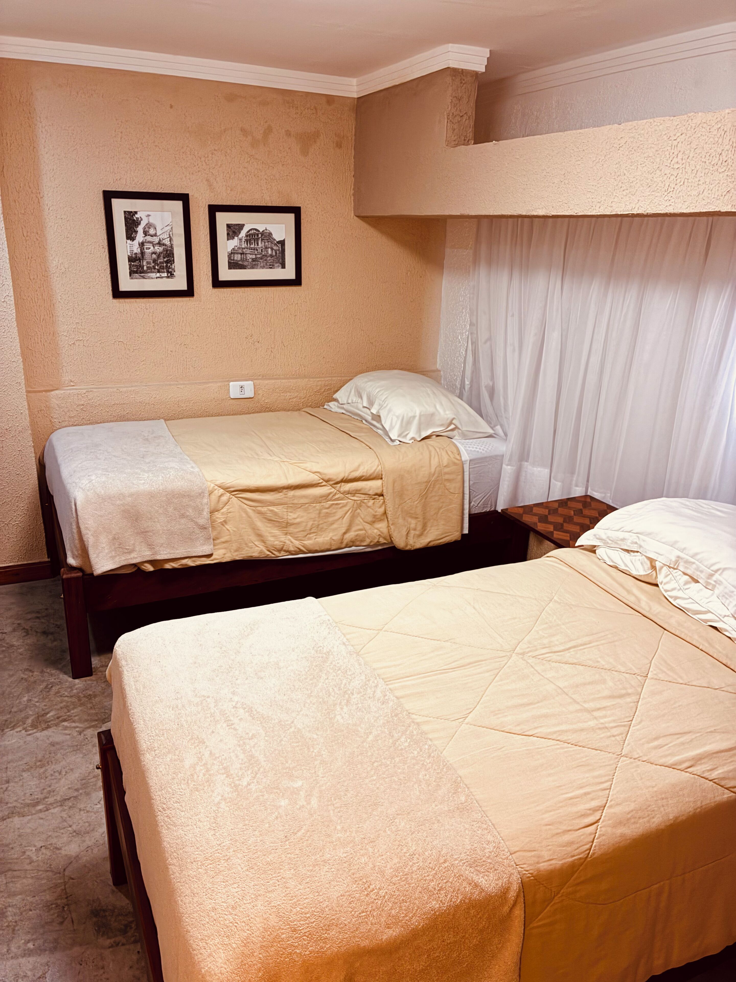 Una habitación con dos camas, una litera, un interruptor de luz en la pared y dos cuadros enmarcados.