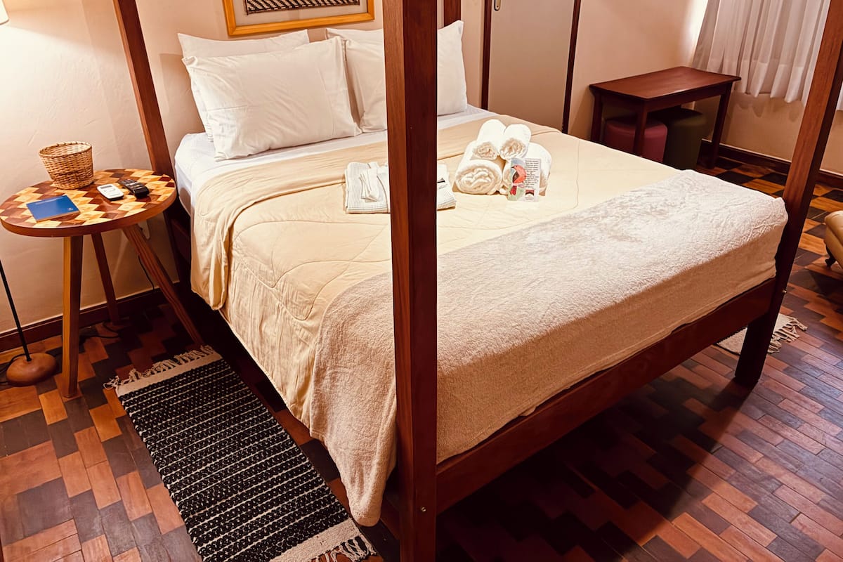 A four-poster bed with a beige bedspread, a bedside table with a basket and a remote control, and a small rug on the floor.