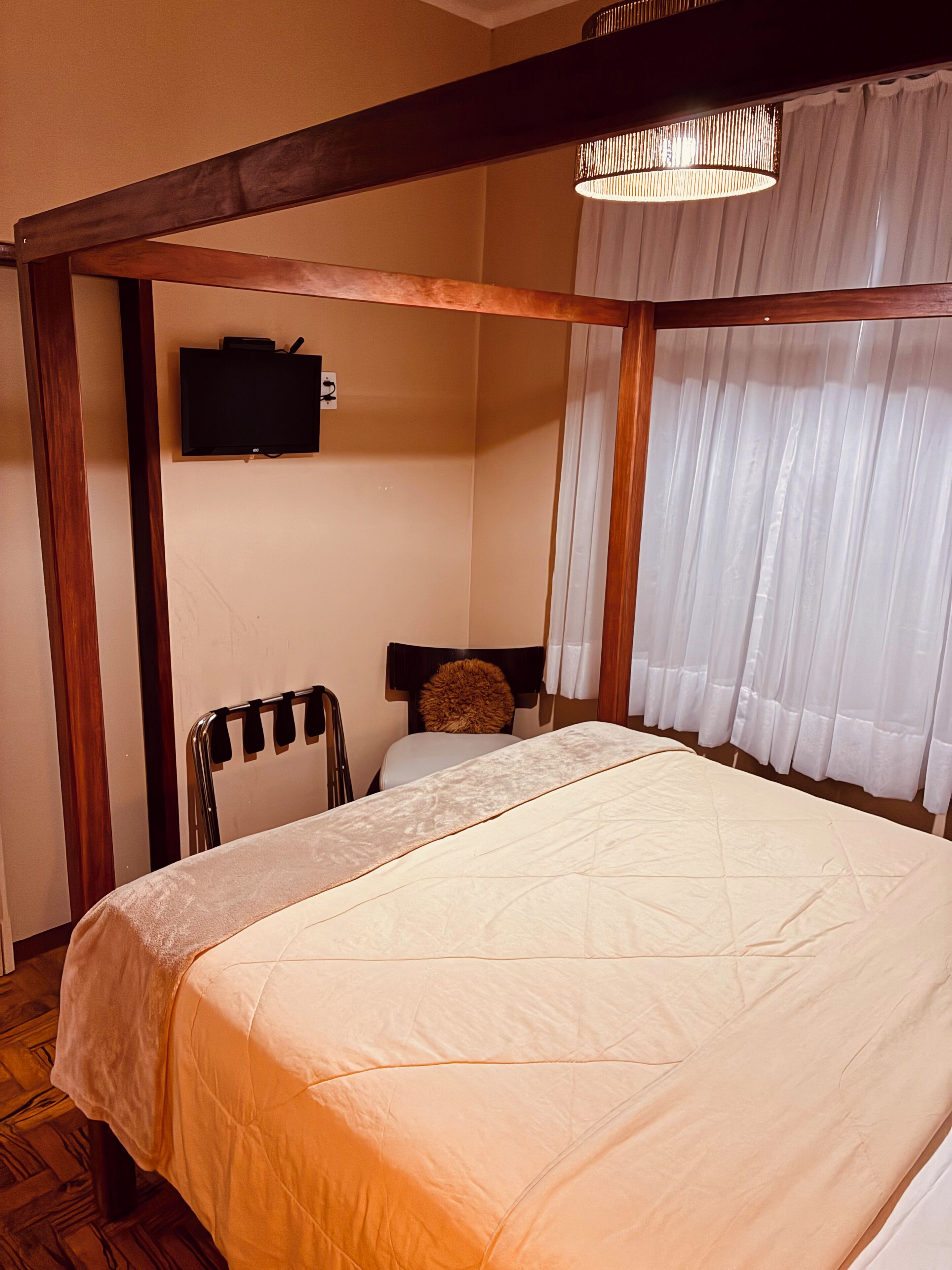Un dormitorio con cama, televisor en la pared, una silla y una ventana con cortinas.