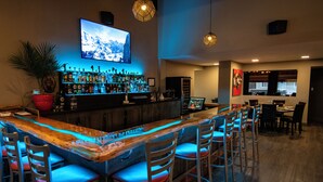 Un espace bar avec un comptoir en bois, de nombreux tabourets, un téléviseur fixé au mur et une variété de bouteilles exposées sur des étagères.