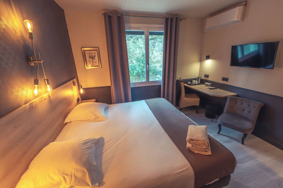 A hotel room with a large bed, a desk, a chair, a TV, and a window with curtains.