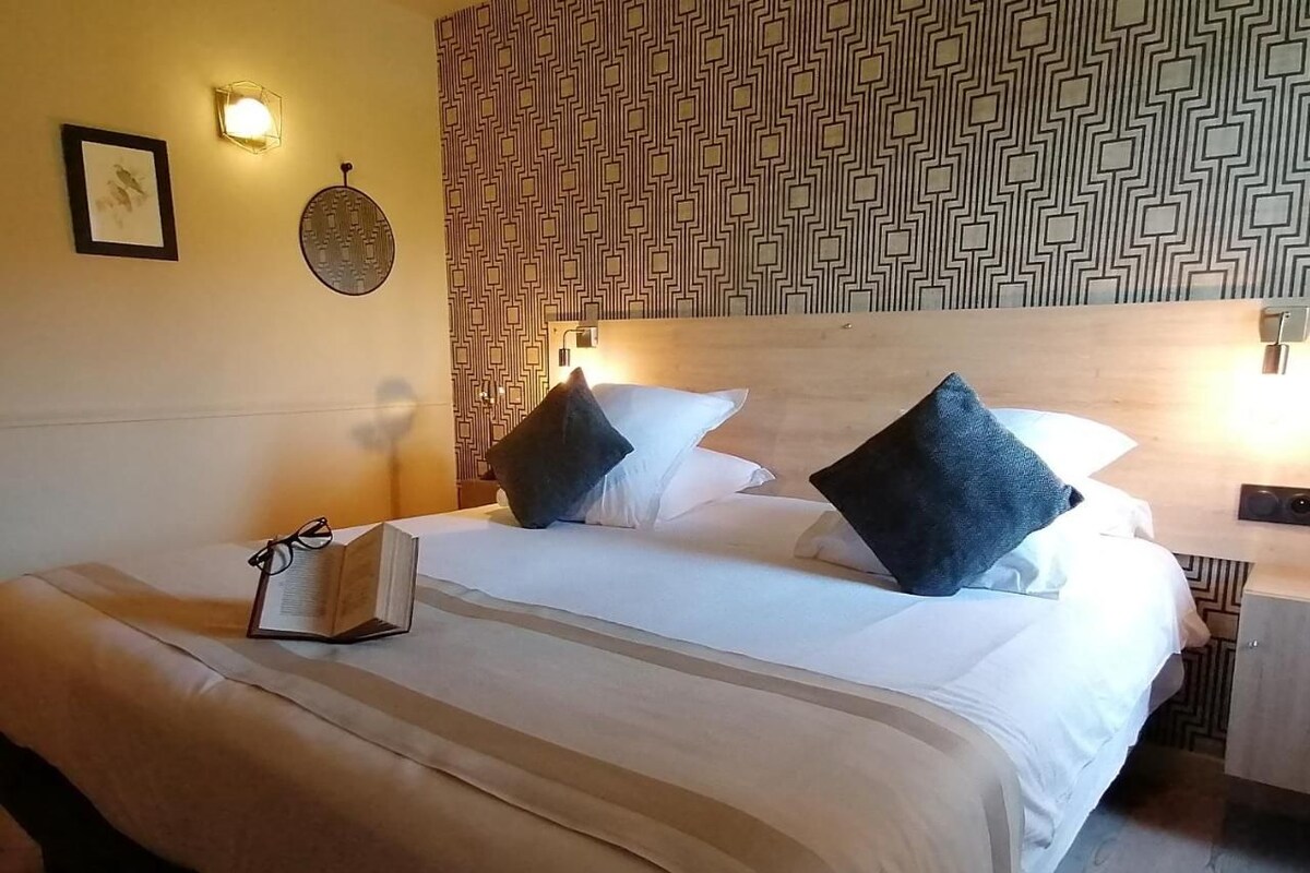 Una cama bien hecha con almohadas, una mesita de noche con una lámpara y un papel tapiz estampado.