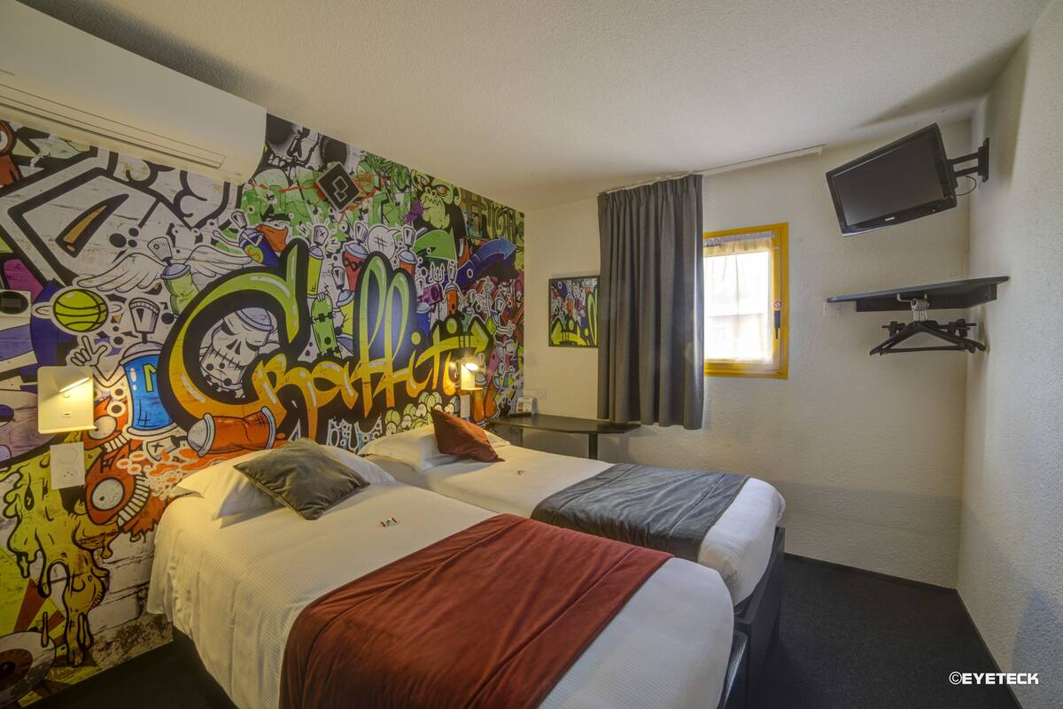 A hotel room with two beds, a mural on the wall, a TV, and a window with curtains.