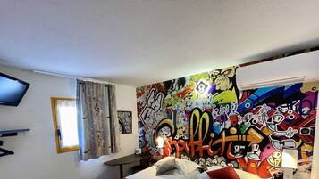 Una habitación con un mural de grafitis en la pared, un televisor de pantalla plana montado, una cama con almohadas y una ventana con cortinas.