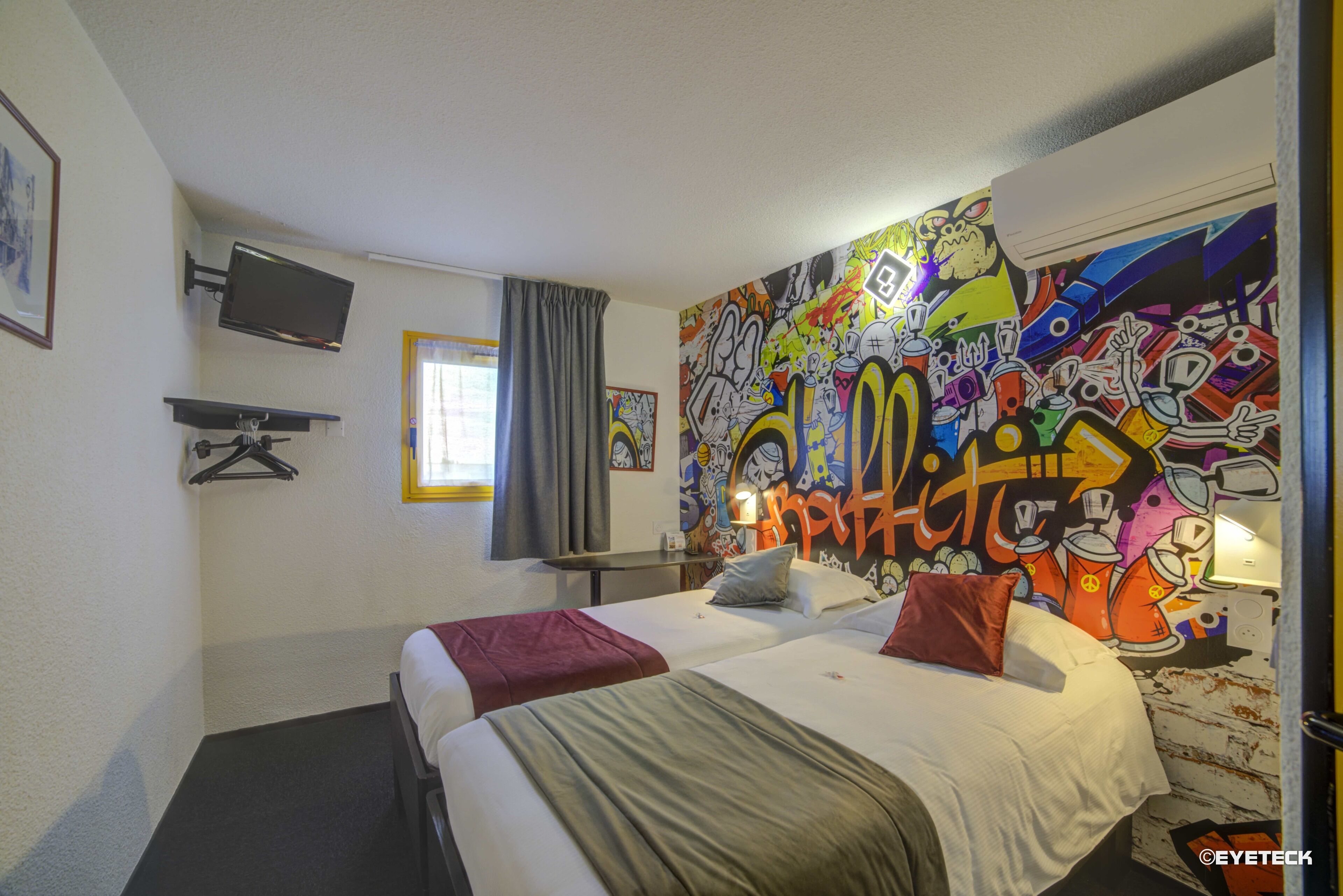 Habitació d'hotel amb dos llits, televisió, finestra amb cortines i una paret coberta de grafitis.