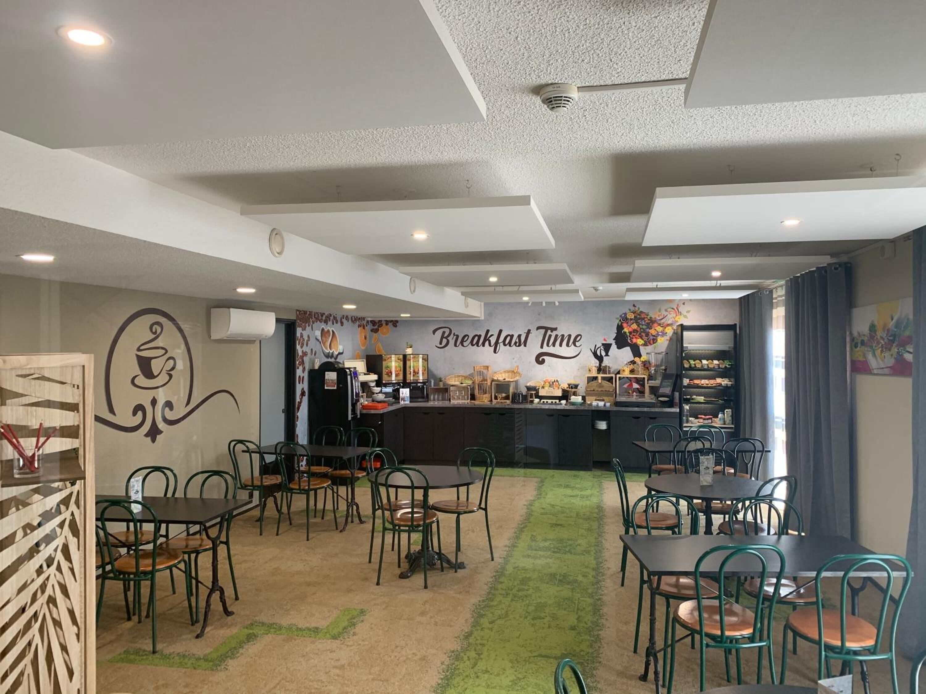 Un área de desayuno con mesas y sillas, una barra con exhibición de alimentos y una pared con un mural que dice 'Breakfast Time'.