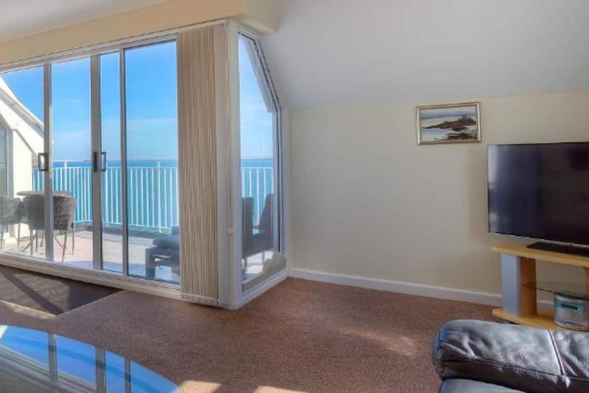 Una sala de estar con un sofá, una mesa de centro de vidrio, un televisor y un balcón con vista al mar.