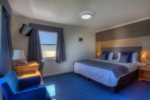 Ein Hotelzimmer mit einem großen Bett, einem Schreibtisch mit Lampe, einem an der Wand befestigten Fernseher und einem Fenster mit Vorhängen.