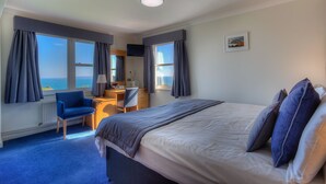 A hotel room with a large bed, a desk, a chair, and a view of the sea. - Hotel Jerbourg (St. Martins)