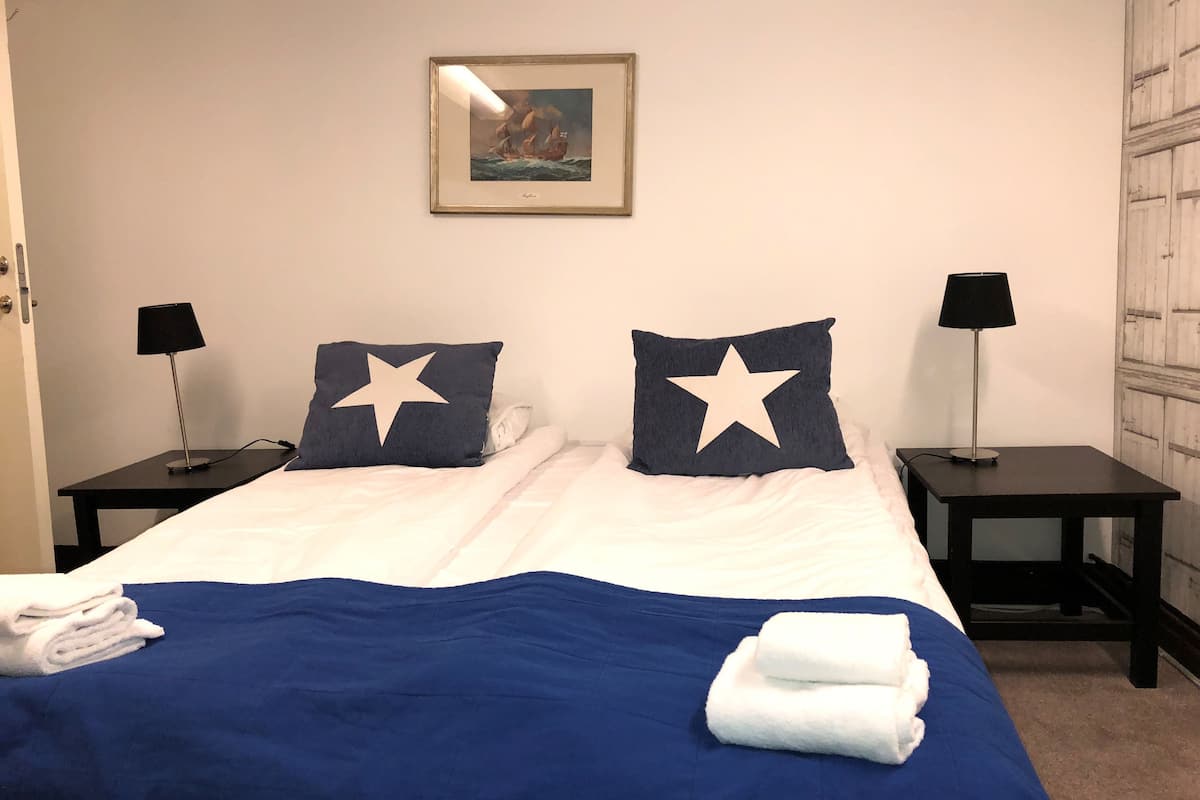 Een net opgemaakt bed met een blauwe spreideken en twee kussens met sterrenmotieven, omringd door twee nachtkastjes met lampen, een ingelijste foto aan de muur, en een deur aan de linkerkant.