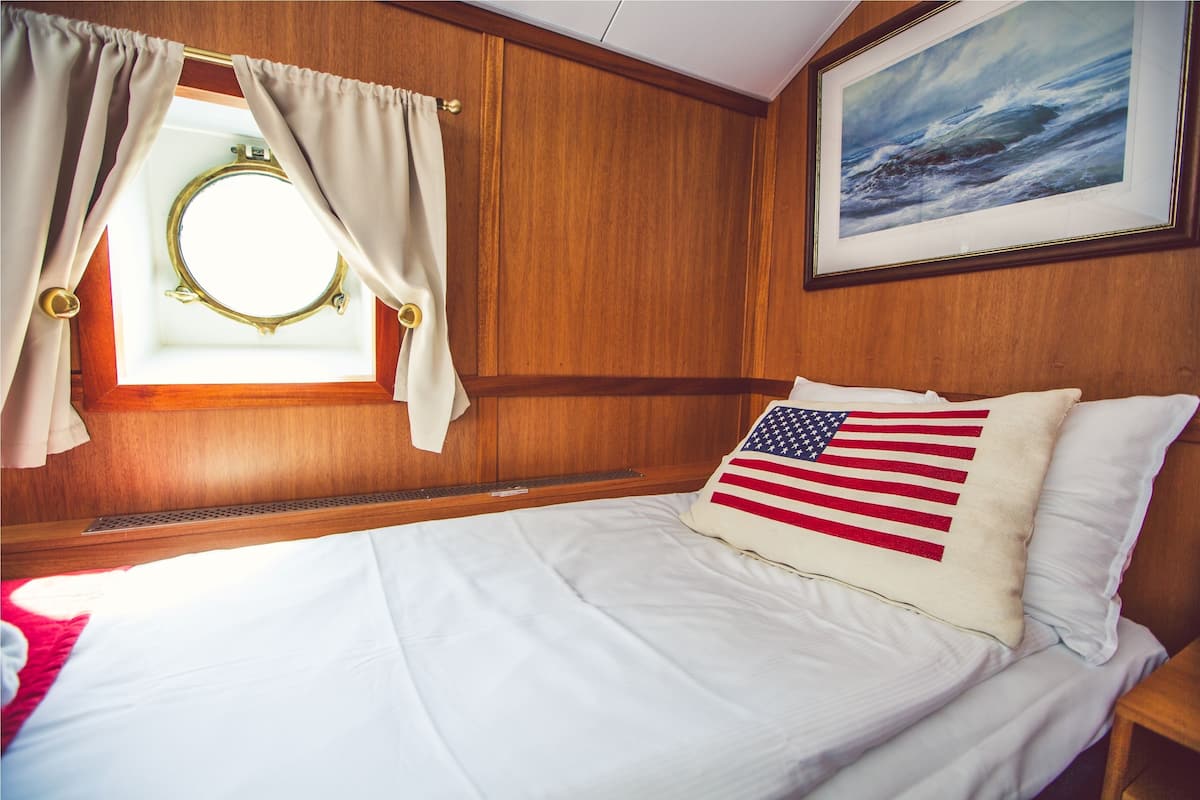 Een hotelkamer met een wand bekleed met houtpanelen, een ronde spiegel, een bed met wit beddengoed, een kussen met de Amerikaanse vlag en een schilderij in een lijst boven het bed.