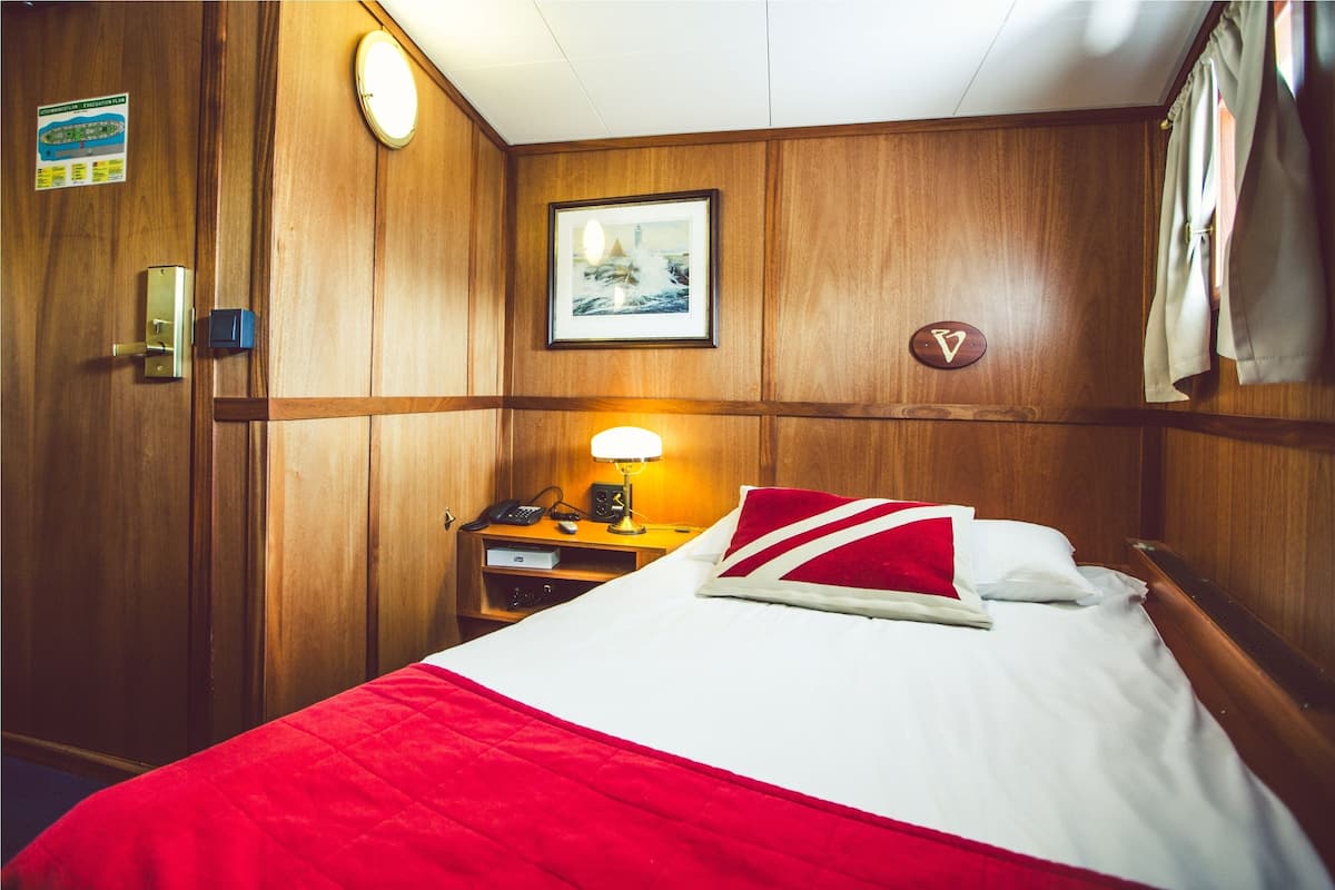 Hotelkamer met een wand van houten panelen, een bed met een rood-witte sprei, een nachtkastje met een lamp, een ingelijst schilderij en een deur met een bordje.