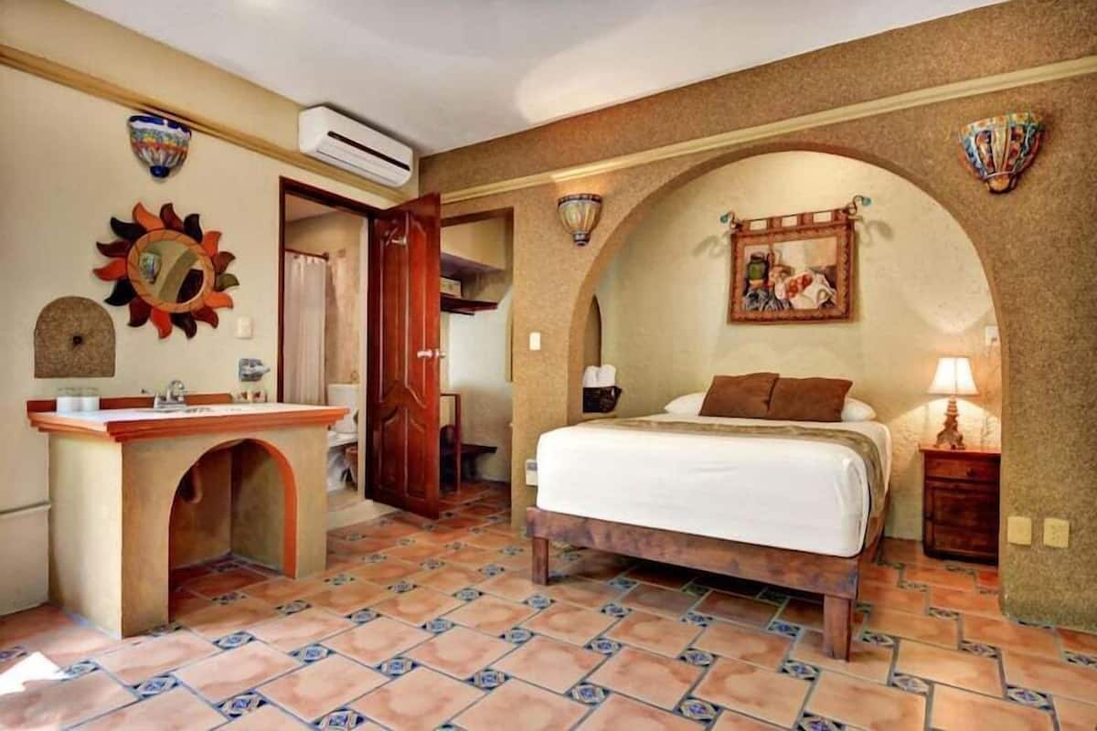A bedroom with a bed, a vanity with a mirror, a wall-mounted heater, and a door leading to another room.