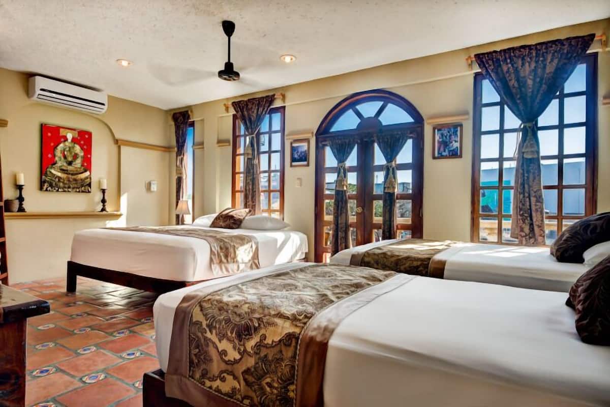 A hotel room with three beds, large windows with curtains, a ceiling fan, and artwork on the walls.