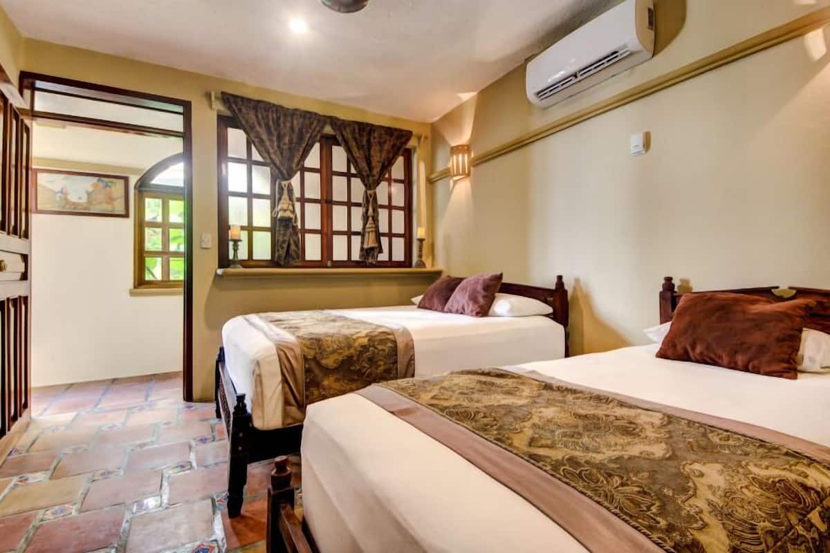 A hotel room with two beds, a patterned rug, a window with curtains, and an air conditioning unit.