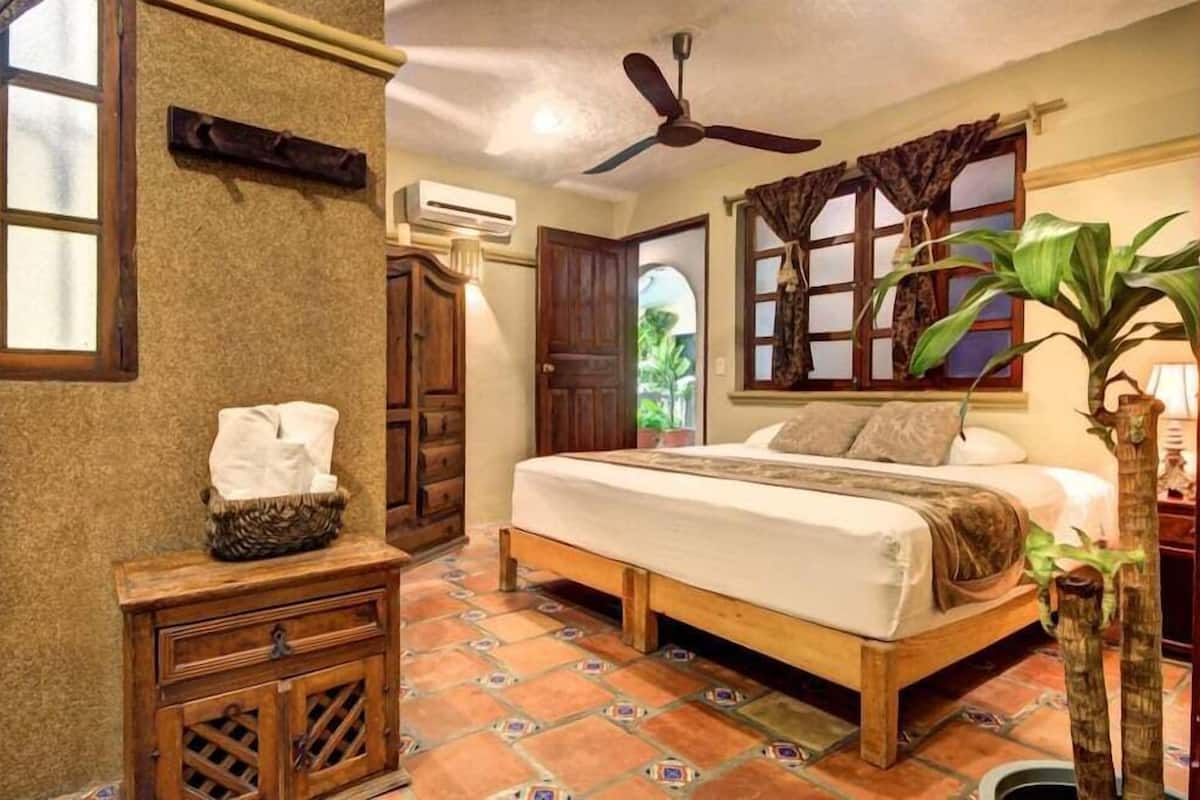 A bedroom with a wooden bed, a ceiling fan, a window with curtains, a potted plant, and a small wooden cabinet.