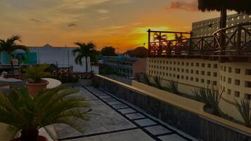 Una terraza en la azotea con vista al atardecer, palmeras y un edificio con techo de paja.