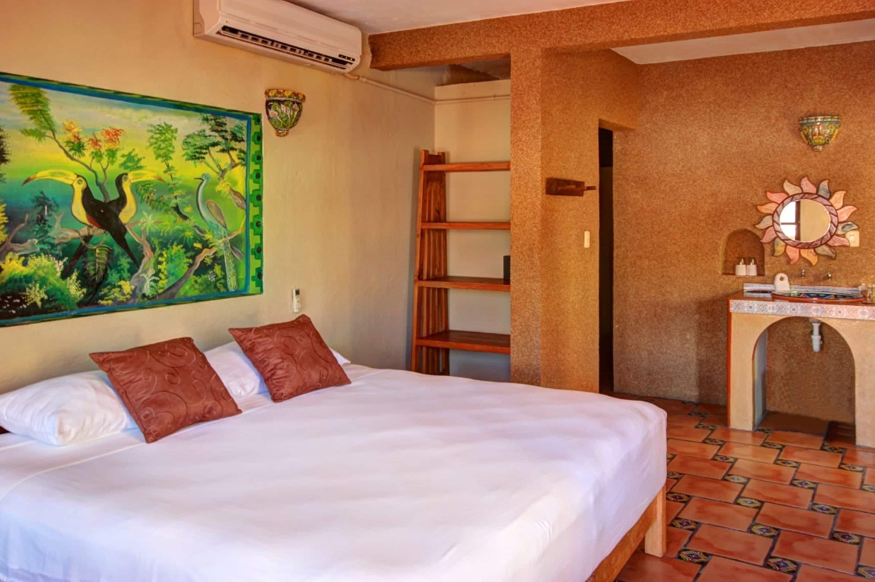 Habitación de hotel con cama, un cuadro de tucanes, estante de madera y baño con espejo y lavamanos.