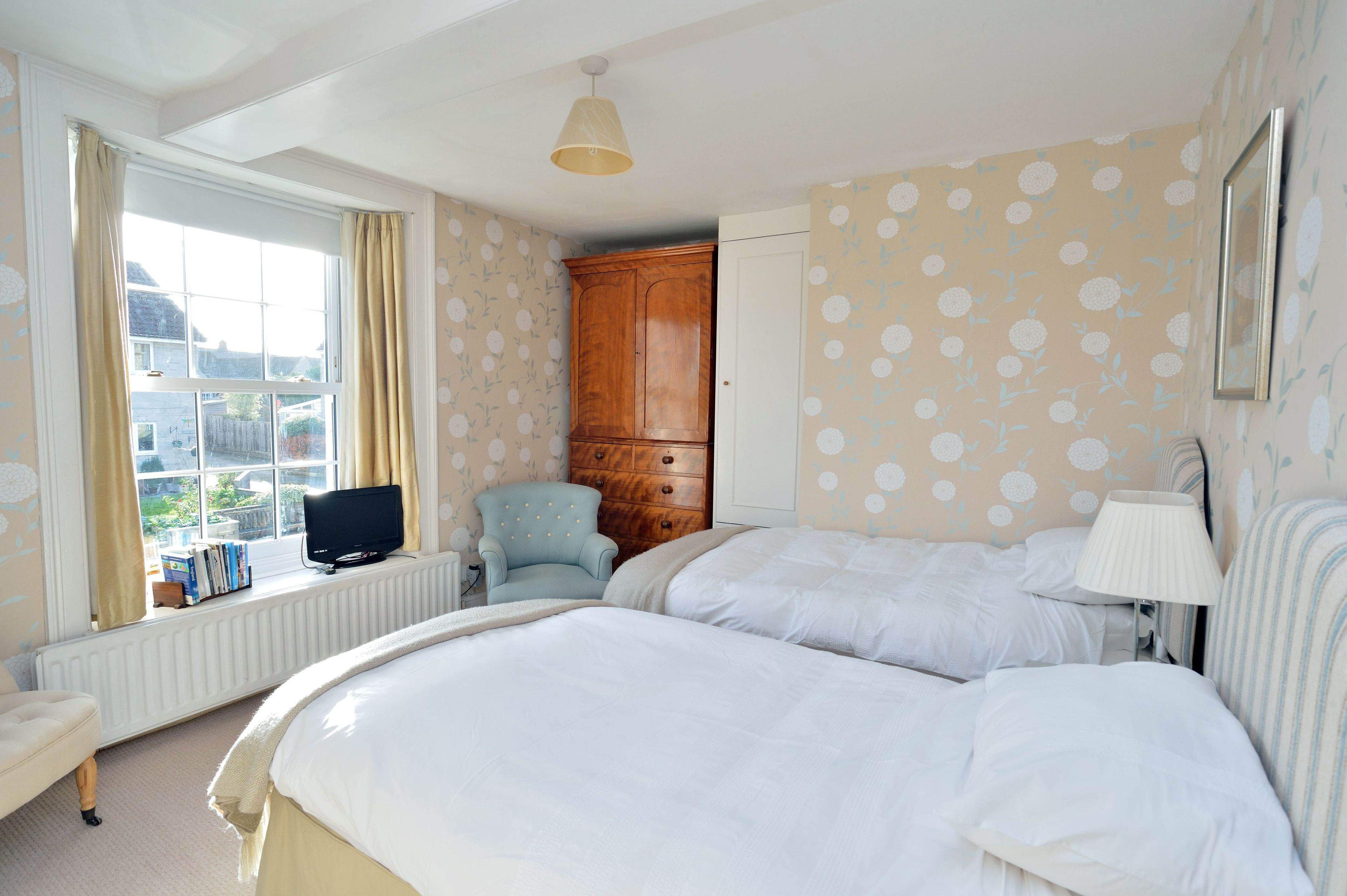 A bedroom with two beds, a wooden wardrobe, a television on a stand, and a window with curtains.