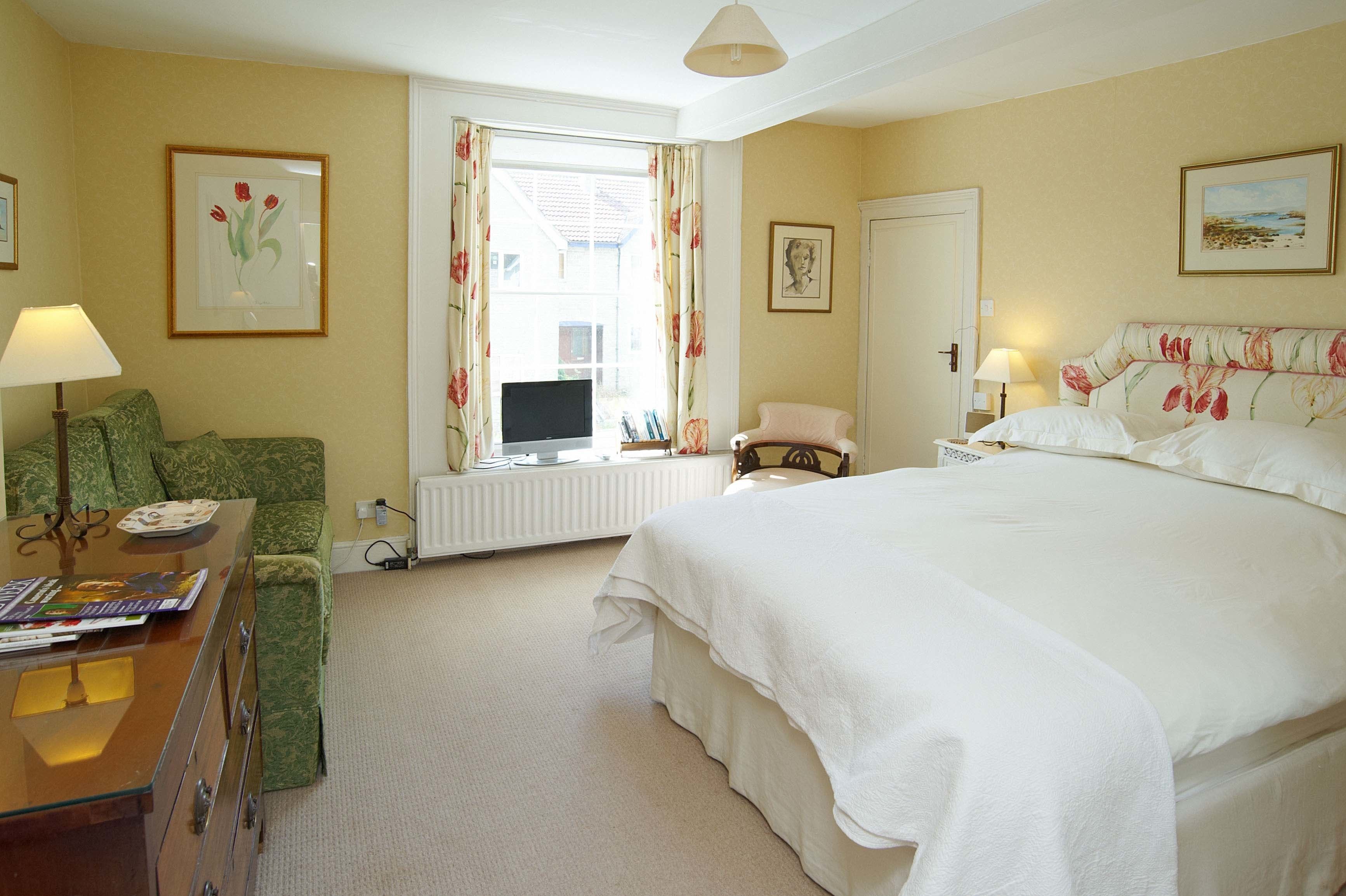 A hotel room with a large bed, a green armchair, a desk with a lamp, and a television.