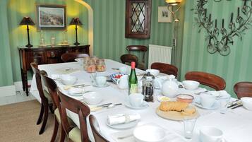 A dining room with a long table set for a meal, surrounded by chairs. There are various dining items on the table, including plates, bowls, and a bottle. The room has green walls and a decorative wall piece.