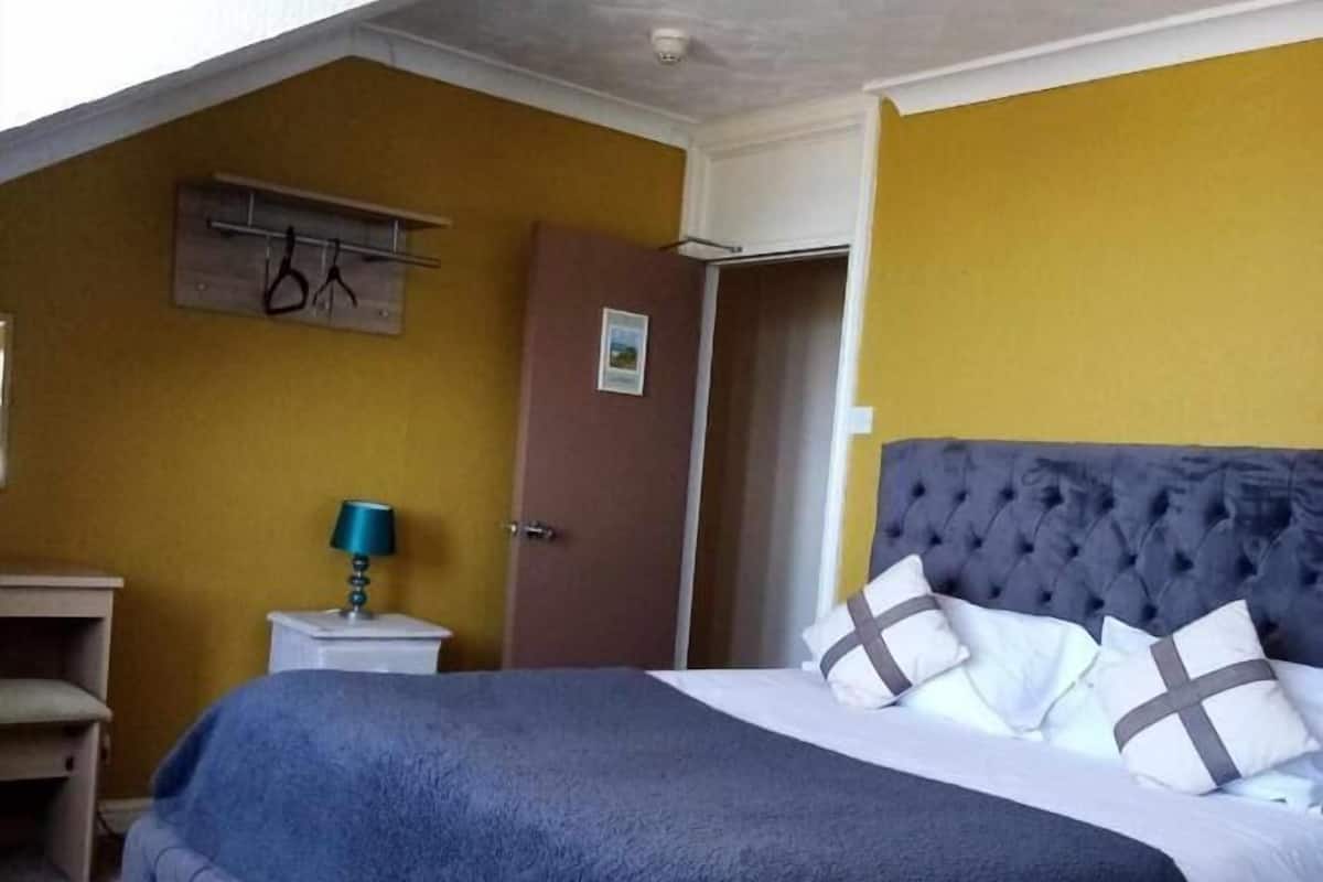 A bedroom with a tufted headboard, a bed with white and gray bedding, a blue blanket, a nightstand with a lamp, and a door with a picture on the wall.