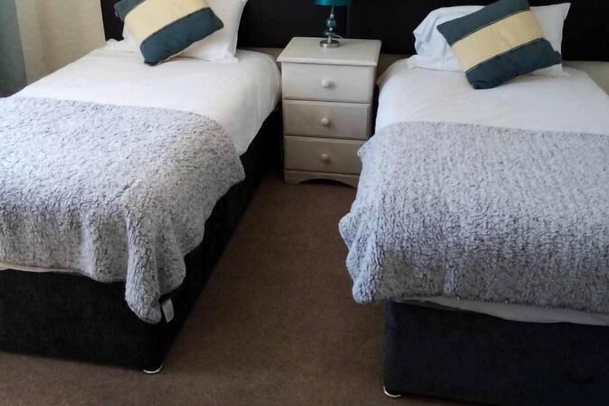 A hotel room with two beds, each with a white blanket and matching pillows. There is a small bedside table with a lamp between the beds.