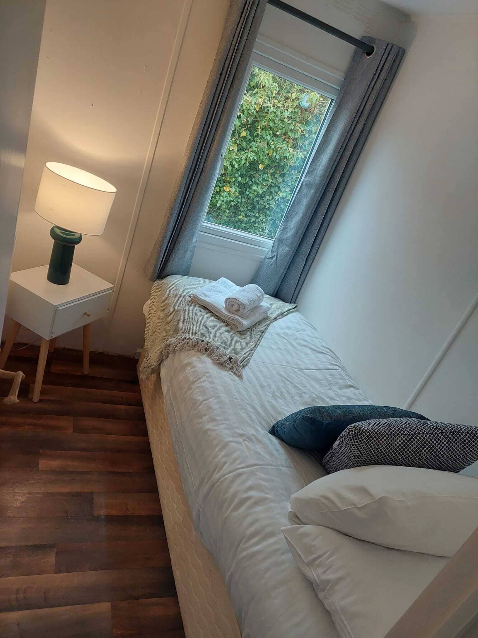 A bedroom with a bed, a nightstand, a lamp, a window with curtains, and a view of greenery outside.