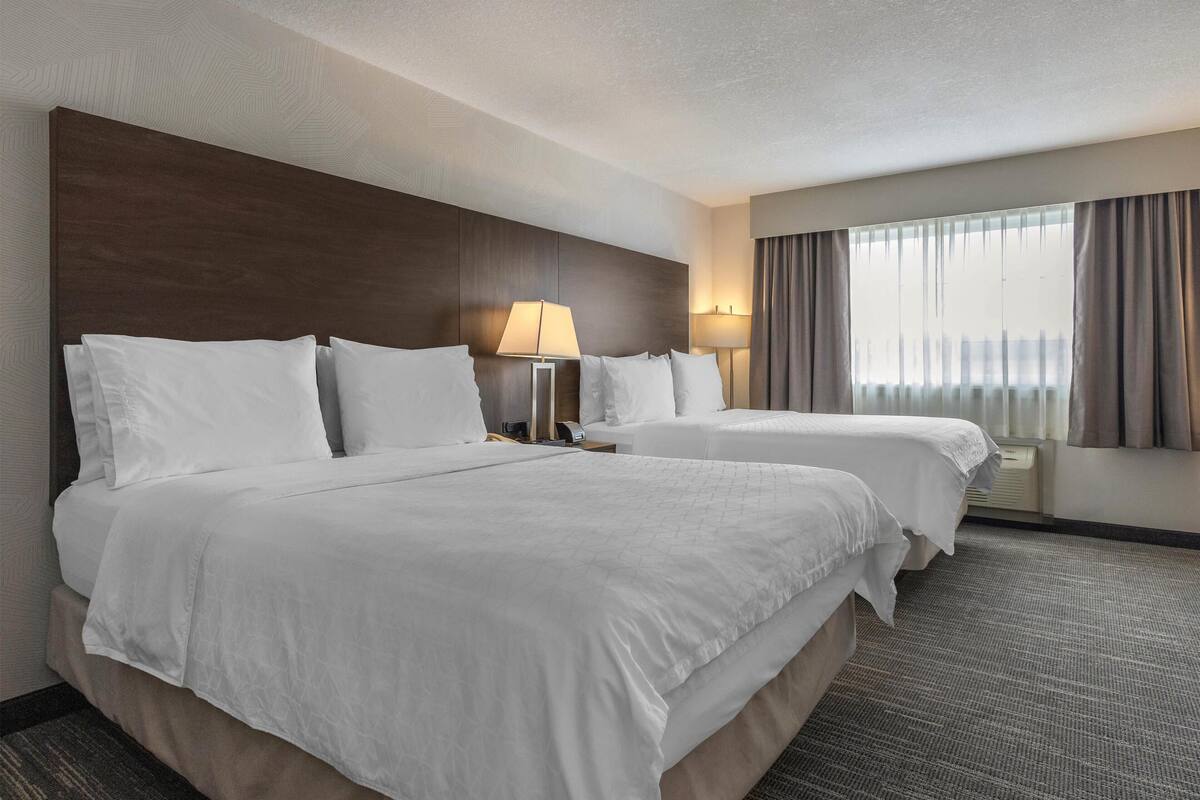 A hotel room with two beds, a large wooden headboard, a window with curtains, and a lamp on each side of the beds.