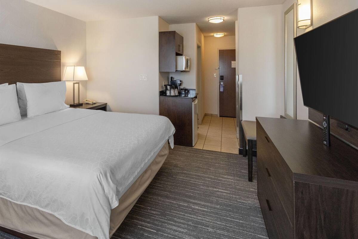 A hotel room with a large bed, a desk with a flat-screen TV, a kitchenette, and a separate bathroom.