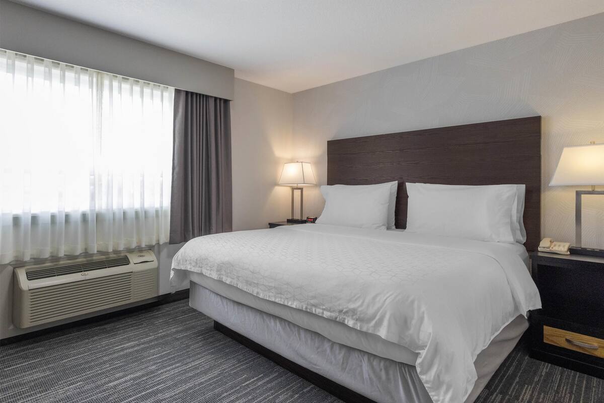 A hotel room with a large bed, a bedside table with a lamp, a wall-mounted air conditioner, and a window with curtains.
