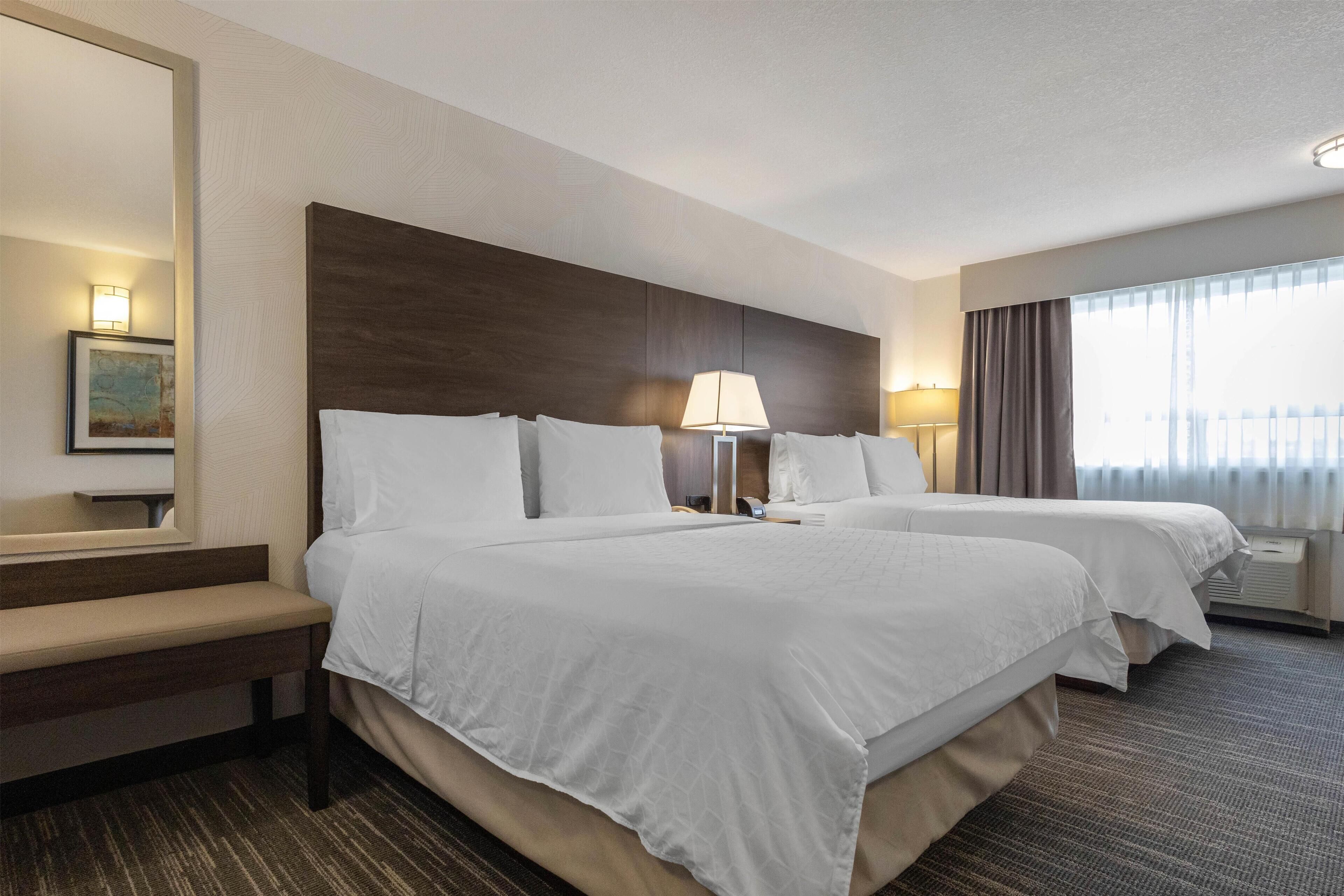 A hotel room with two beds, a bench, a large mirror, and a painting on the wall.