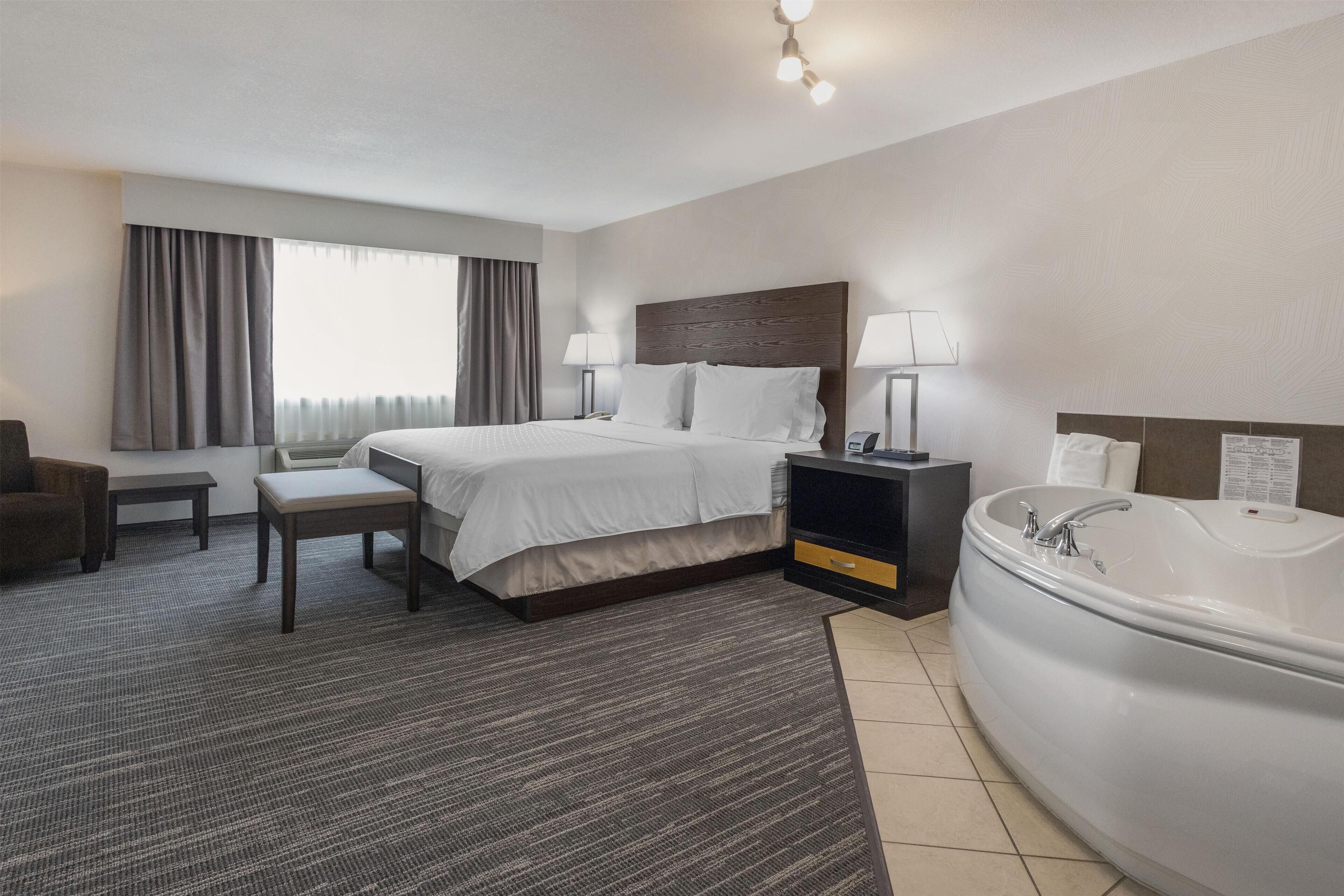A hotel room with a large bed, bedside tables, a desk, a chair, a window with curtains, and a bathtub.