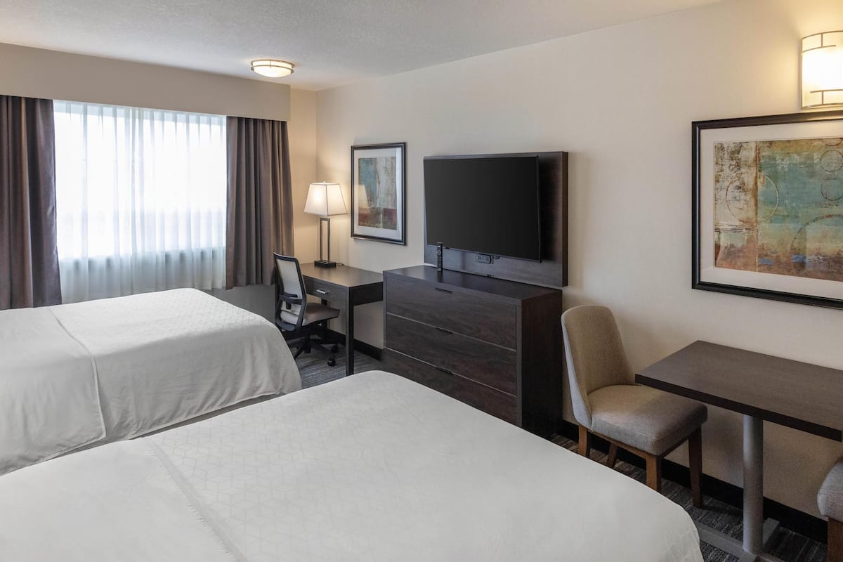 A hotel room with two beds, a desk, a chair, a television, and artwork on the walls.