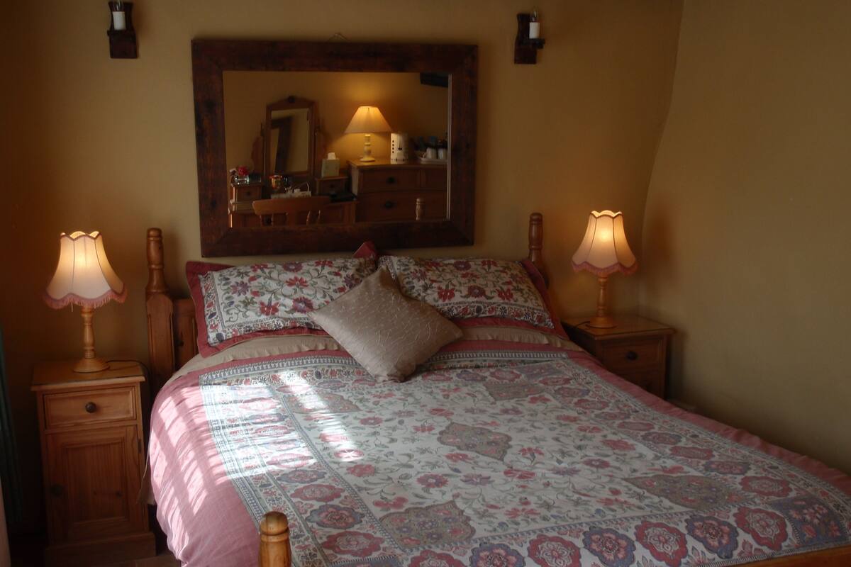 A bedroom with a wooden bed, floral bedding, two bedside lamps, a mirror above the bed, and a nightstand with a lamp.