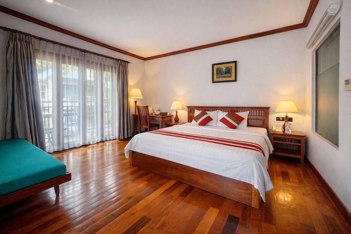 Une chambre d’hôtel avec un plancher en bois, un lit avec une literie blanche et des oreillers rouges et blancs, un bureau avec une chaise, une table de chevet avec une lampe et une fenêtre avec des rideaux.