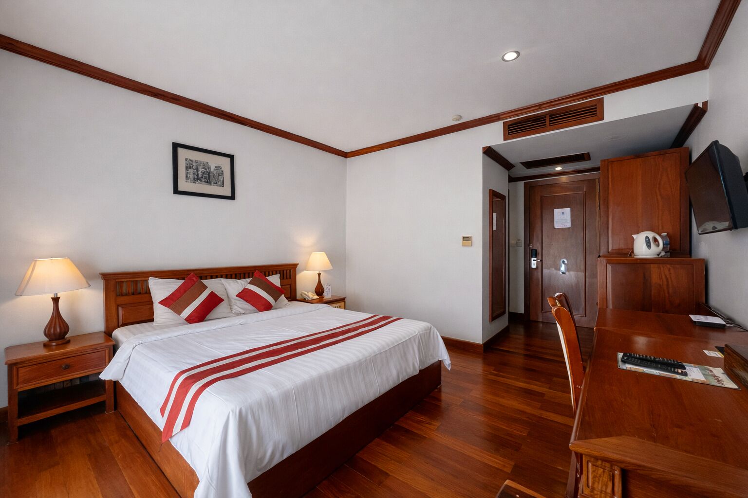 A hotel room with a wooden bed, two bedside lamps, a desk with a chair, and a television on a wooden cabinet.
