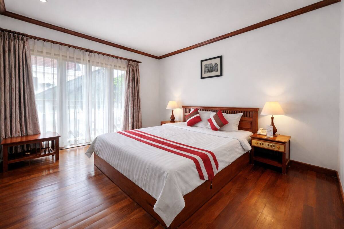 Une chambre d’hôtel avec un grand lit, une tête de lit en bois, deux tables de chevet avec des lampes, un petit banc en bois et une fenêtre avec des rideaux.