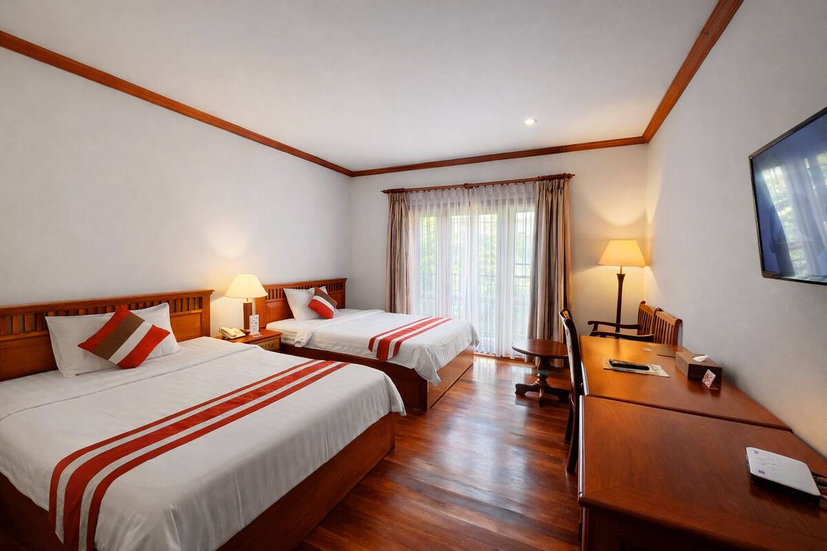 Une chambre d’hôtel avec deux lits, un bureau, une chaise, une lampe et une grande fenêtre avec des rideaux.