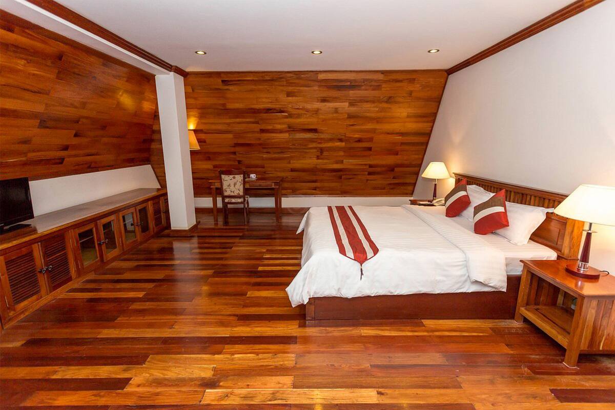 Une chambre avec un plancher en bois, un lit avec une literie blanche et des touches de rouge, une table de chevet avec une lampe et un téléviseur posé sur une commode en bois.