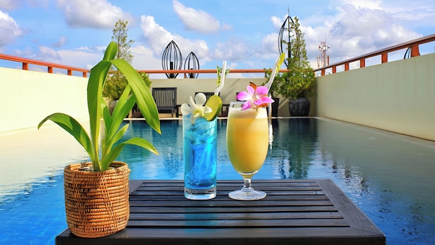 A poolside table with a woven plant pot, a tall glass with blue liquid, and a tall glass with a yellow drink, set against a pool and a building with a red railing. - Cheathata CTA Hotel Siem Reap (Siem Reap)