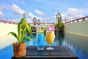 A poolside table with a woven plant pot, a tall glass with blue liquid, and a tall glass with a yellow drink, set against a pool and a building with a red railing. - Cheathata CTA Hotel Siem Reap (Siem Reap)