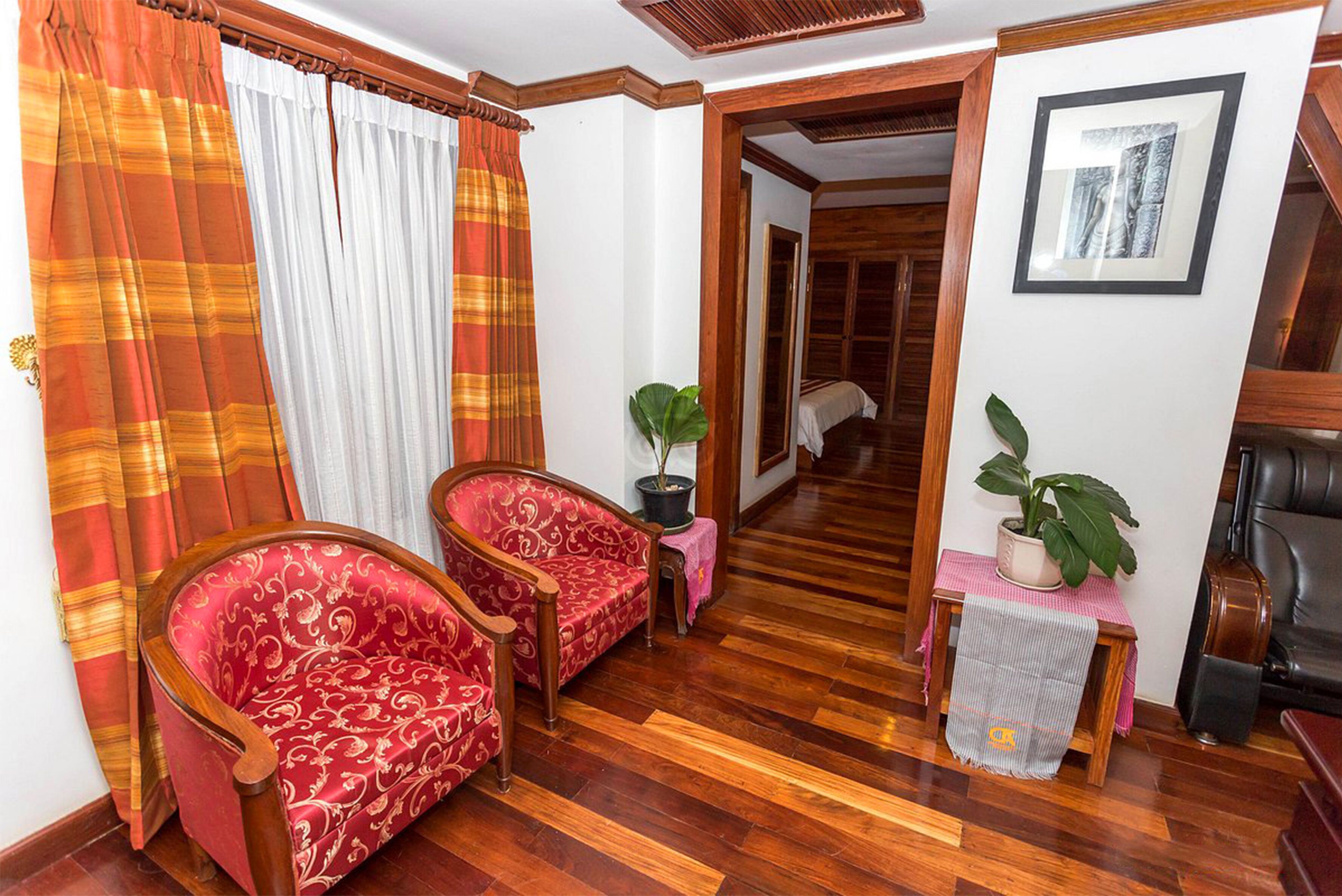 A room with wooden flooring, two red patterned chairs, a small table with a potted plant, and a doorway leading to another room.
