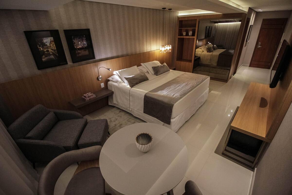 A modern hotel room with a bed, bedside tables, a seating area with a chair and table, a TV, and framed pictures on the wall.
